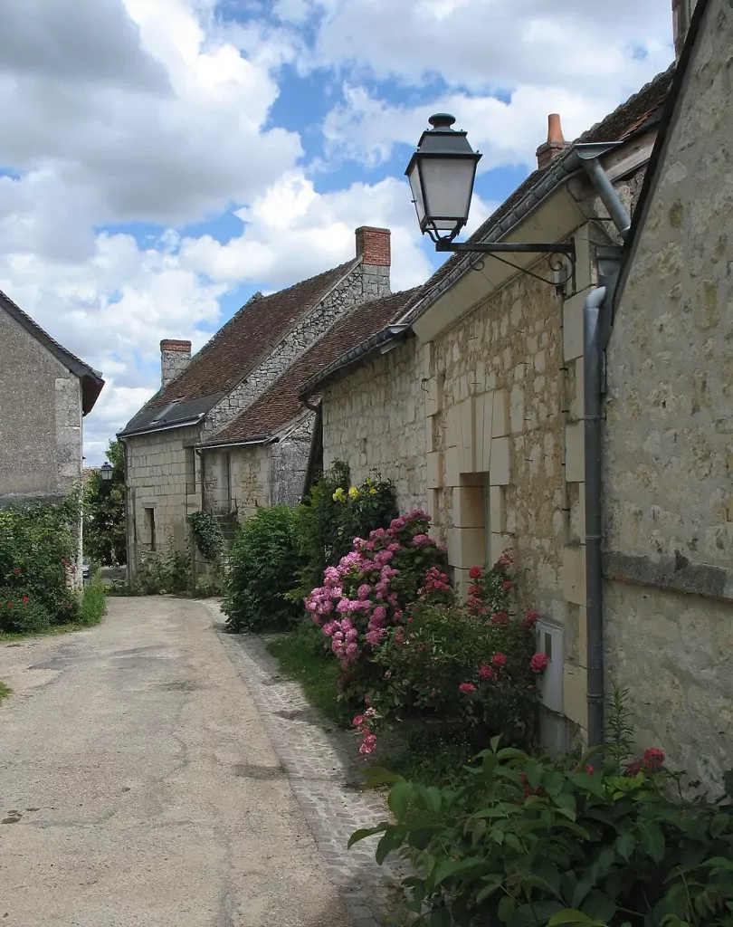 Callejuela empedrada en Crissay-sur-Manse, Francia, con casas de piedra tradicionales y un jardín de rosas bajo un cielo azul con nubes.
