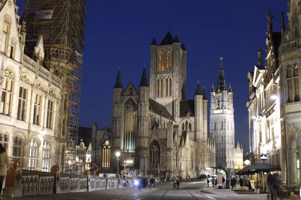 Vista nocturna de Gante con la iglesia de San Nicolás iluminada y la ciudad vibrante en el fondo.