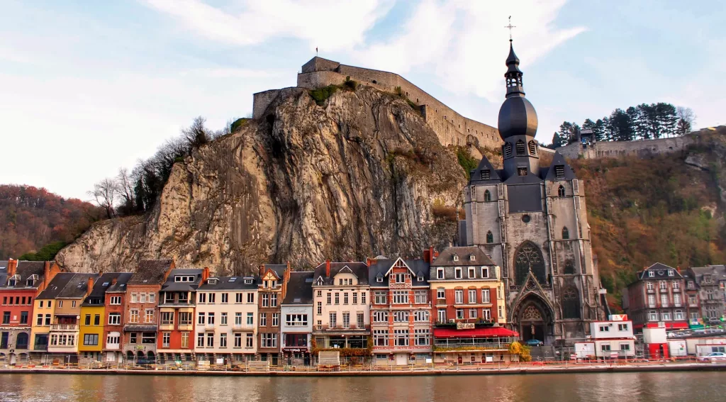 Vista de la ciudad de Dinant, Bélgica, con casas coloridas a orillas del río y el castillo en lo alto de la colina.